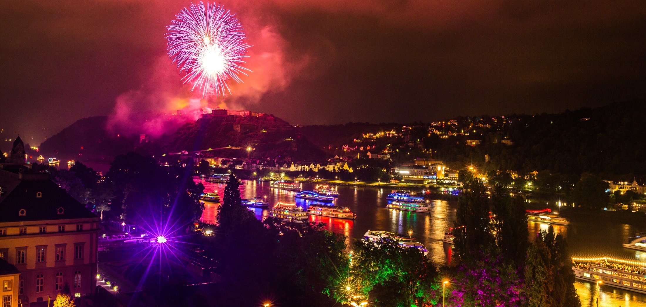 Blick im Dunkeln über die Rheinanlagen auf beleuchtete Schiffe auf dem Rhein und Feuerwerk über der Festung Ehrenbreitstein Blick im Dunkeln über die Rheinanlagen auf beleuchtete Schiffe auf dem Rhein und Feuerwerk über der Festung Ehrenbreitstein