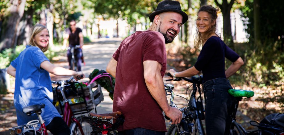 Radfahrende in Grünanlage - zu sehen: zwei Frauen und ein Mann auf ihren Fahrrädern, mit fröhlichem Blick in Richtung Kamera