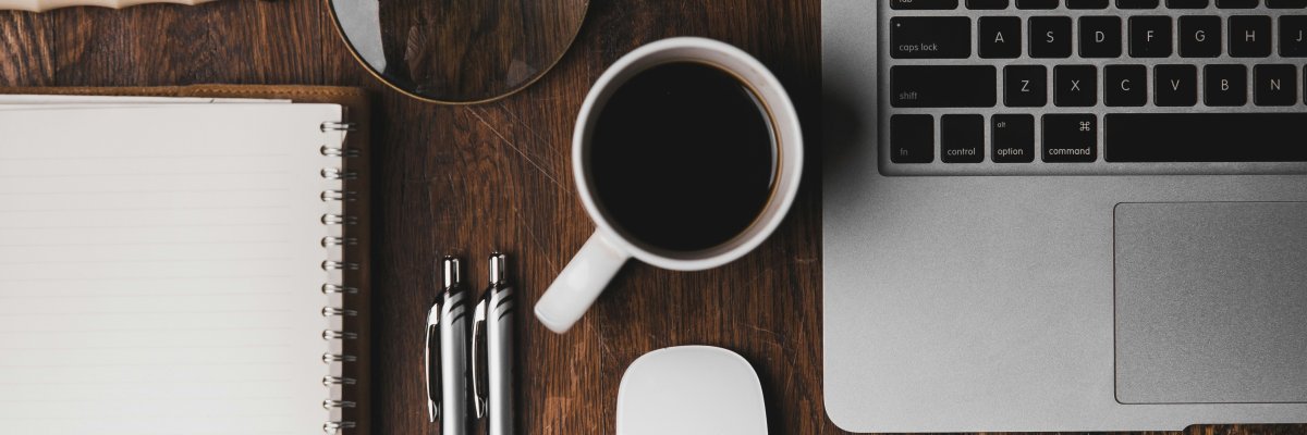 Ein Schreibtisch mit Tasse, Laptop, Schreibblock und zwei Kugelschreibern