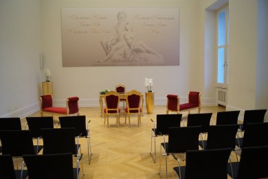 Blick in den Augusta-Saal des kurfürstlichen Schloss. Zu sehen sind die Stuhlreihen der Gäste des Brauptpaares sowie der goldene Trautisch mit Sitzmöglichkeiten für das Brautpaar und deren Trauezeugen sowie den Standesbeamten. Auf der Hinteren Wand ist ein Foto von "Vater Rhein" und "Mutter Mosel" zu sehen.