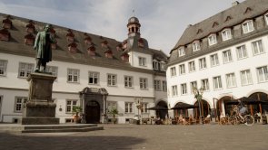 Jesuitenplatz mit zwei weißen Gebäuden, Platzfläche und Denkmal darauf