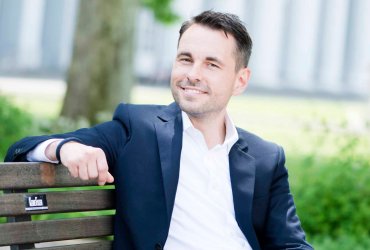 Oberbürgermeister David Langner - auf Bank sitzend, blaues Sakko, weißes Hemd, Jeans, im Hintergrund ein Baum