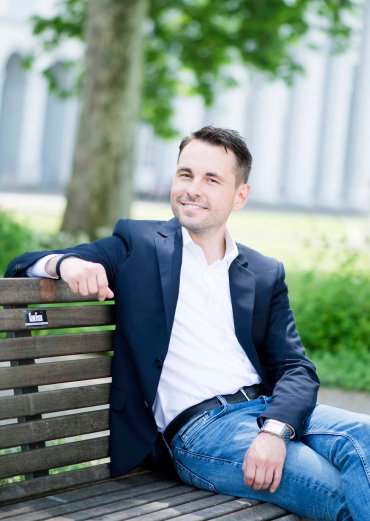 Oberbürgermeister David Langner - auf Bank sitzend, blaues Sakko, weißes Hemd, Jeans, im Hintergrund ein Baum