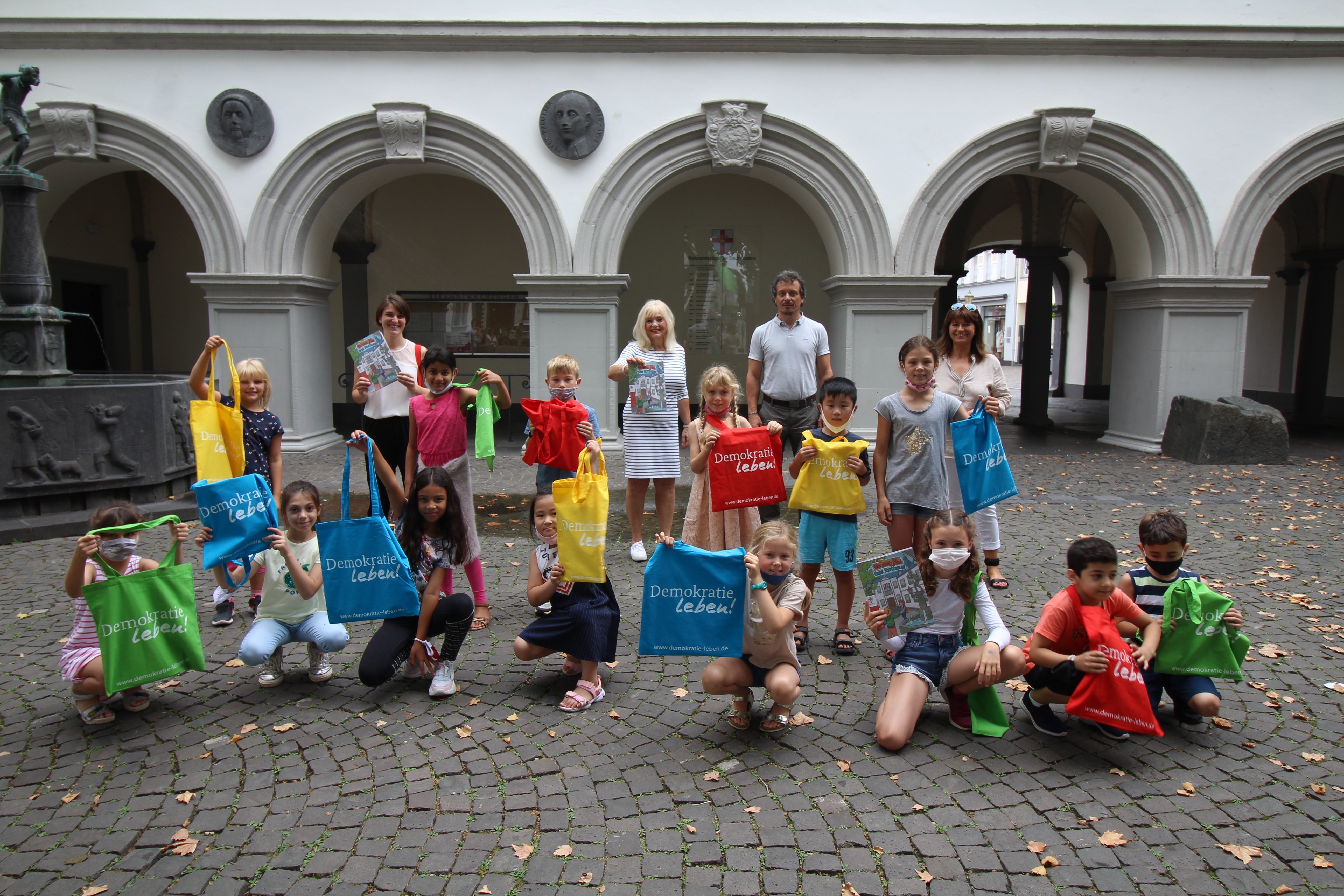 Grundschulkinder erhalten Lernmalhefte zum Thema Kommunalpolitik. Grundschulkinder halten Lernmalhefte und bunte Taschen hoch, die sie von der Dezernentin für Bildung und Kultur überreicht bekommen haben. Die Hefte thematisieren Kommunalpolitik.