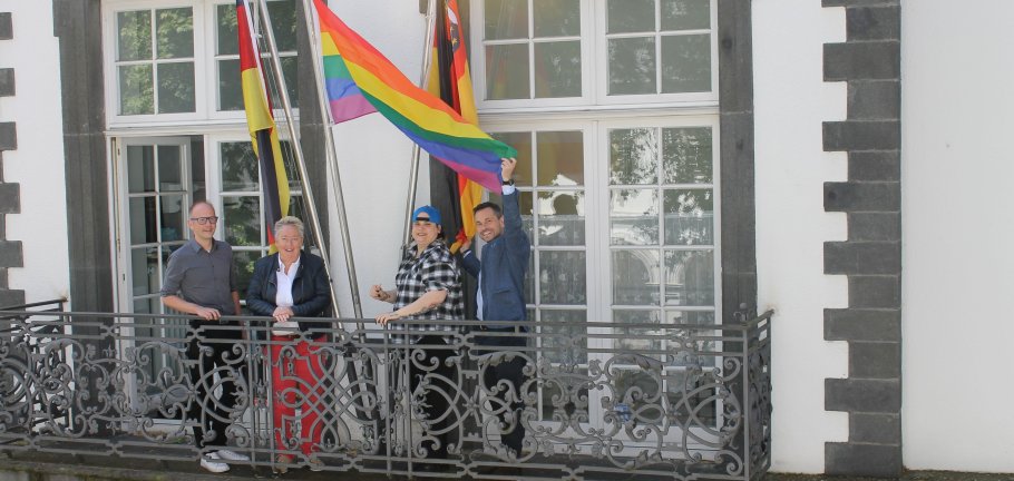 Vier Personen stehen auf einem Balkon und hantieren an einer Regenbogenfahne.
