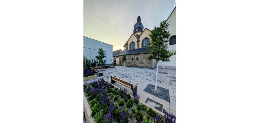 Ein neu angelegter Platz, im Vordergrund Blumenbeete. Im Hintergrund eine Kirche.