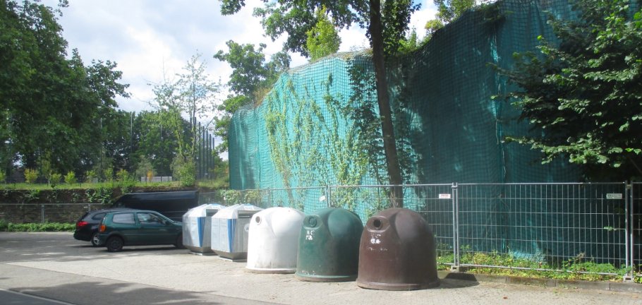 5 Container stehen vor einem Bauzaun. Im Hintergrund parken Autos.