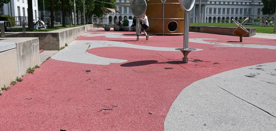 Ein Speilplatz mit beschädigtem Bodenbelag. Im Hintergrund ein spielendes Kind an einem Gerät.