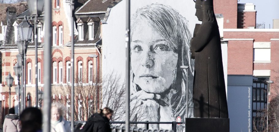 Auf einer Hauswand das riesige Portrait einer jungen Frau. Im Vordergrund Menschen auf einer Brücke.