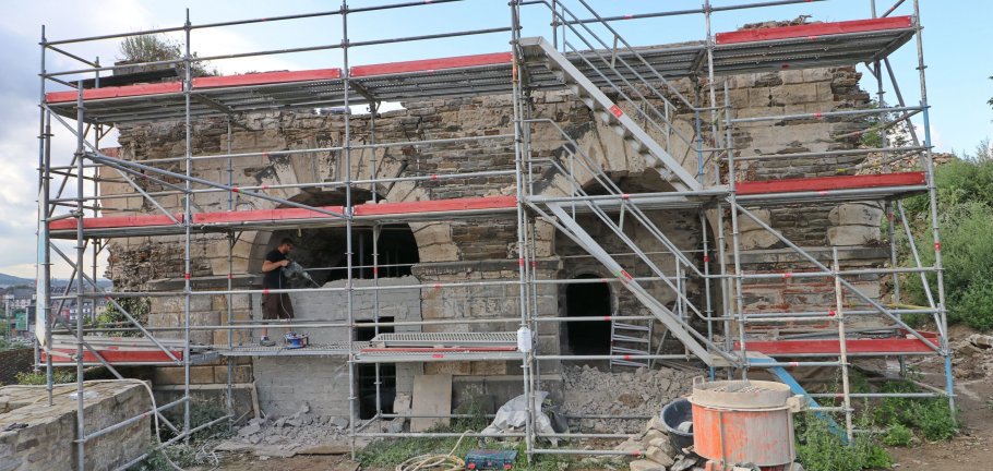 Hinter einem Gerüst, auf dem ein Arbeiter steht, zwei rundbögige Eingänge in ein altes Gemäuer.