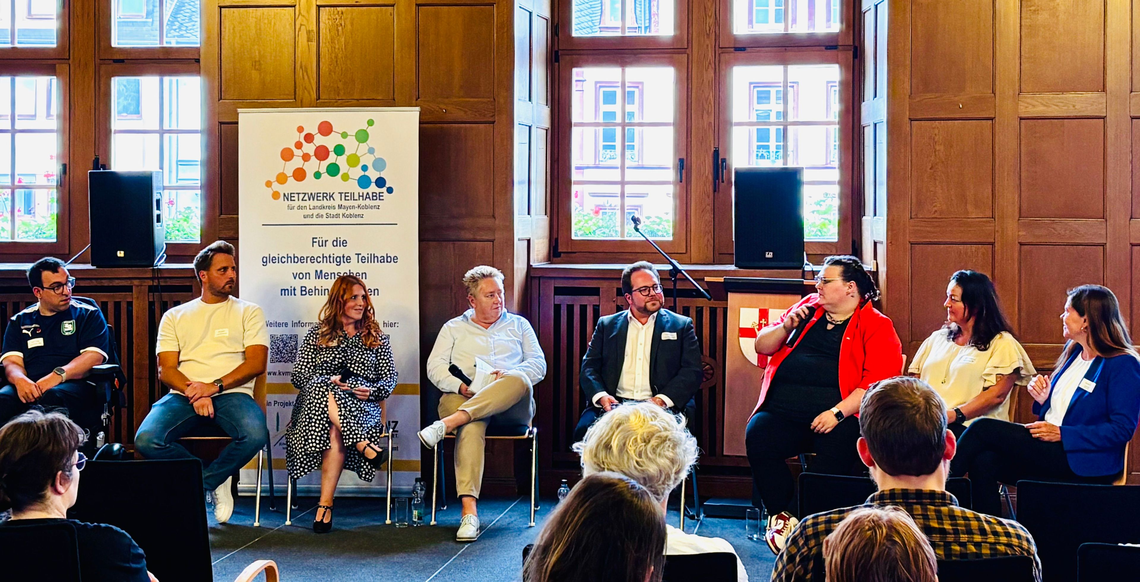 Viele Menschen auf einem Podium in eine Diskussion vertieft. Im Vordergrund die Hinterköpfe von Zuschauern.