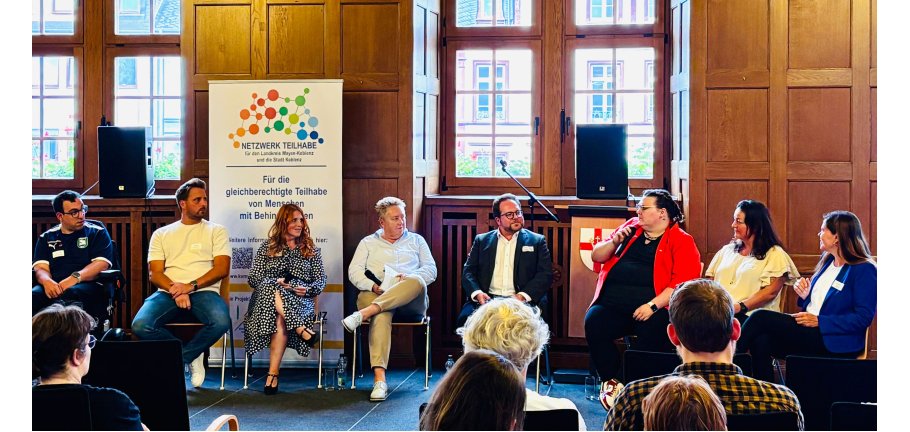 Viele Menschen auf einem Podium in eine Diskussion vertieft. Im Vordergrund die Hinterköpfe von Zuschauern.