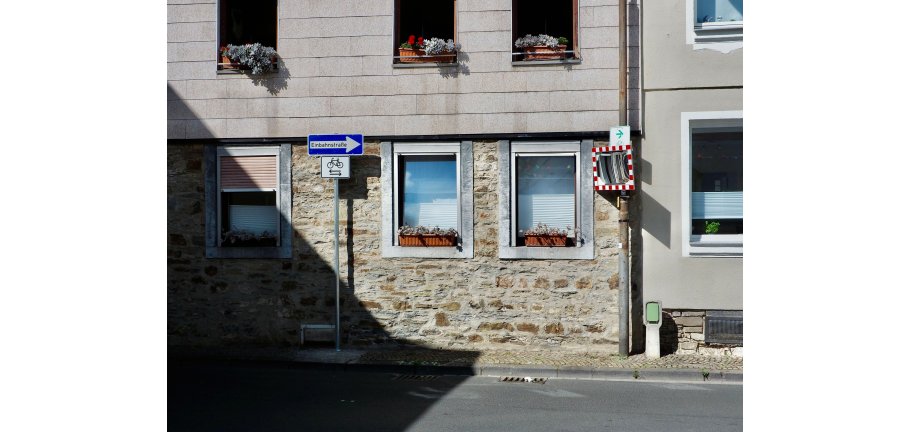 Blick über eine STraße auf Hauswände vor denen ein Verkehrsspiegel und ein Verkehrsschild stehen.