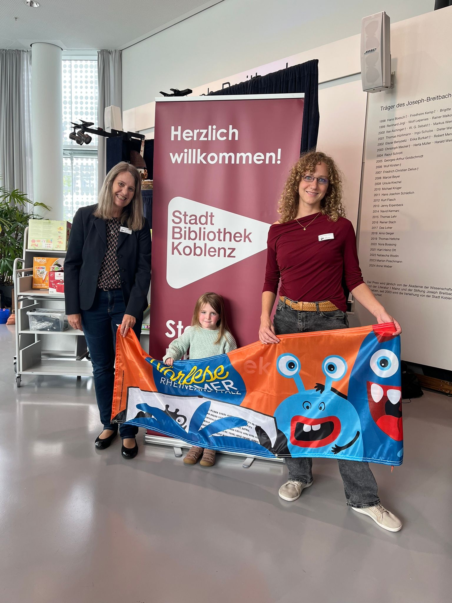 Zwei erwachsene Frauen umrahmen ein kleines Kind. Alle drei halten ein Banner des Vorlese-Sommers.