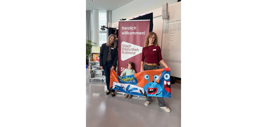 Zwei erwachsene Frauen umrahmen ein kleines Kind. Alle drei halten ein Banner des Vorlese-Sommers.