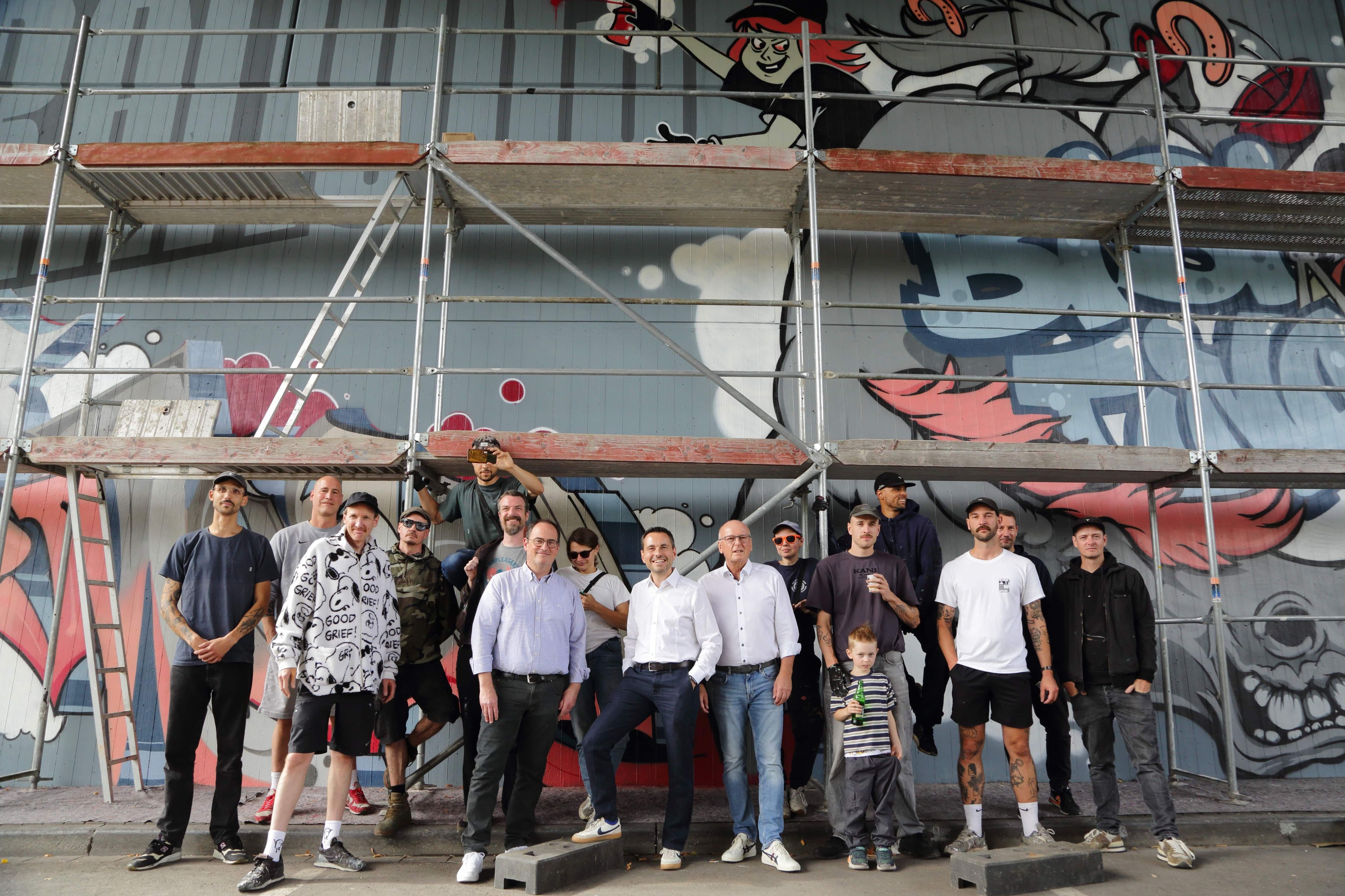 Eine Gruppe von Menschen steht vor einem Brückenpfeiler vor dem ein Gerüst steht. Der Pfeiler ist bemalt.