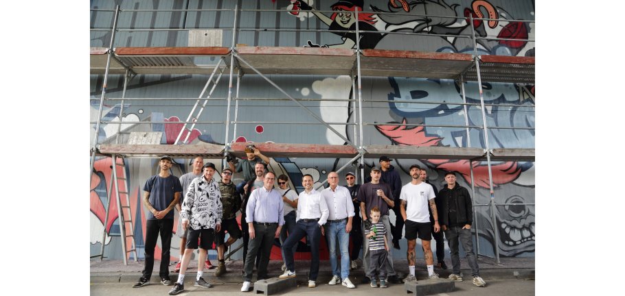 Eine Gruppe von Menschen steht vor einem Brückenpfeiler vor dem ein Gerüst steht. Der Pfeiler ist bemalt.