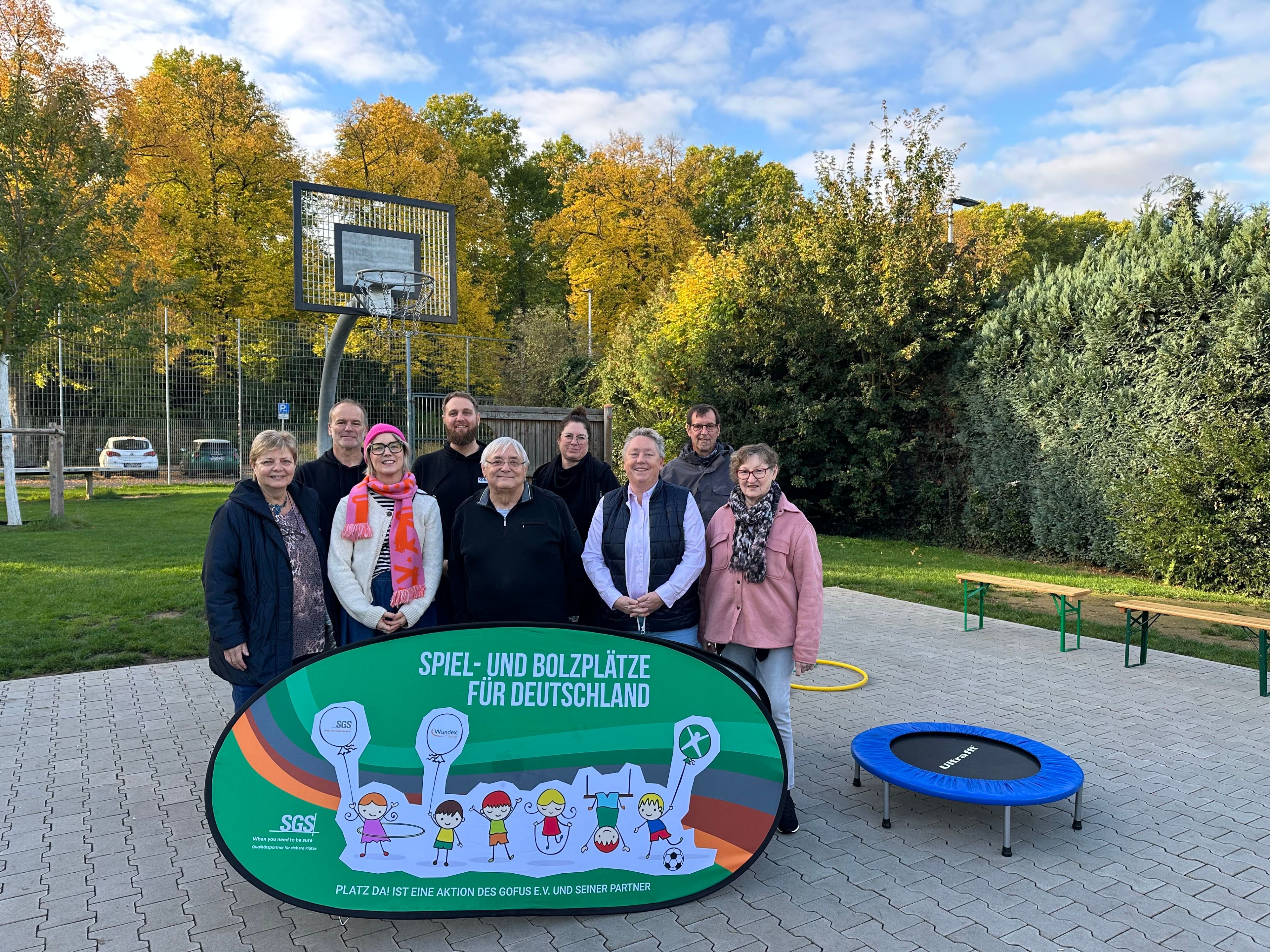 Viele Personen stehen hinter einem bunten Plakat. Im Hintergrund Spiel- und Sportgeräte.