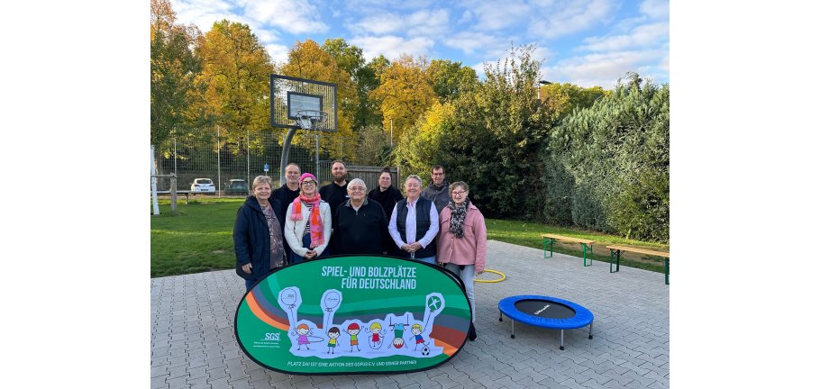 Viele Personen stehen hinter einem bunten Plakat. Im Hintergrund Spiel- und Sportgeräte.