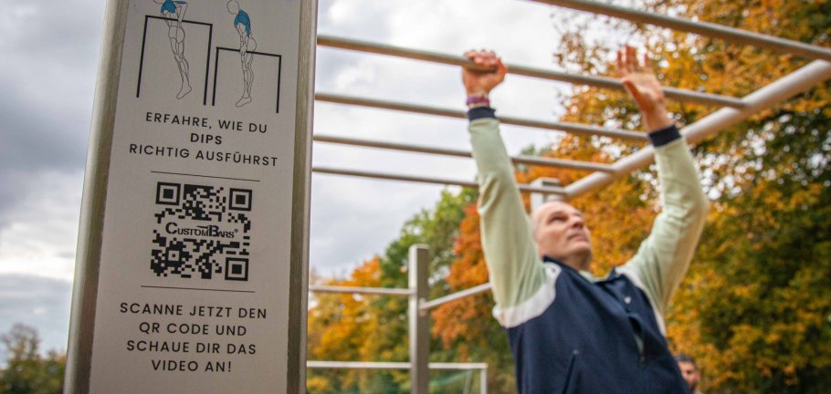 Ein QR-Code ist im Vordergrund zu sehen, während im Hintergrund sich an Stangen entlang hangelt.