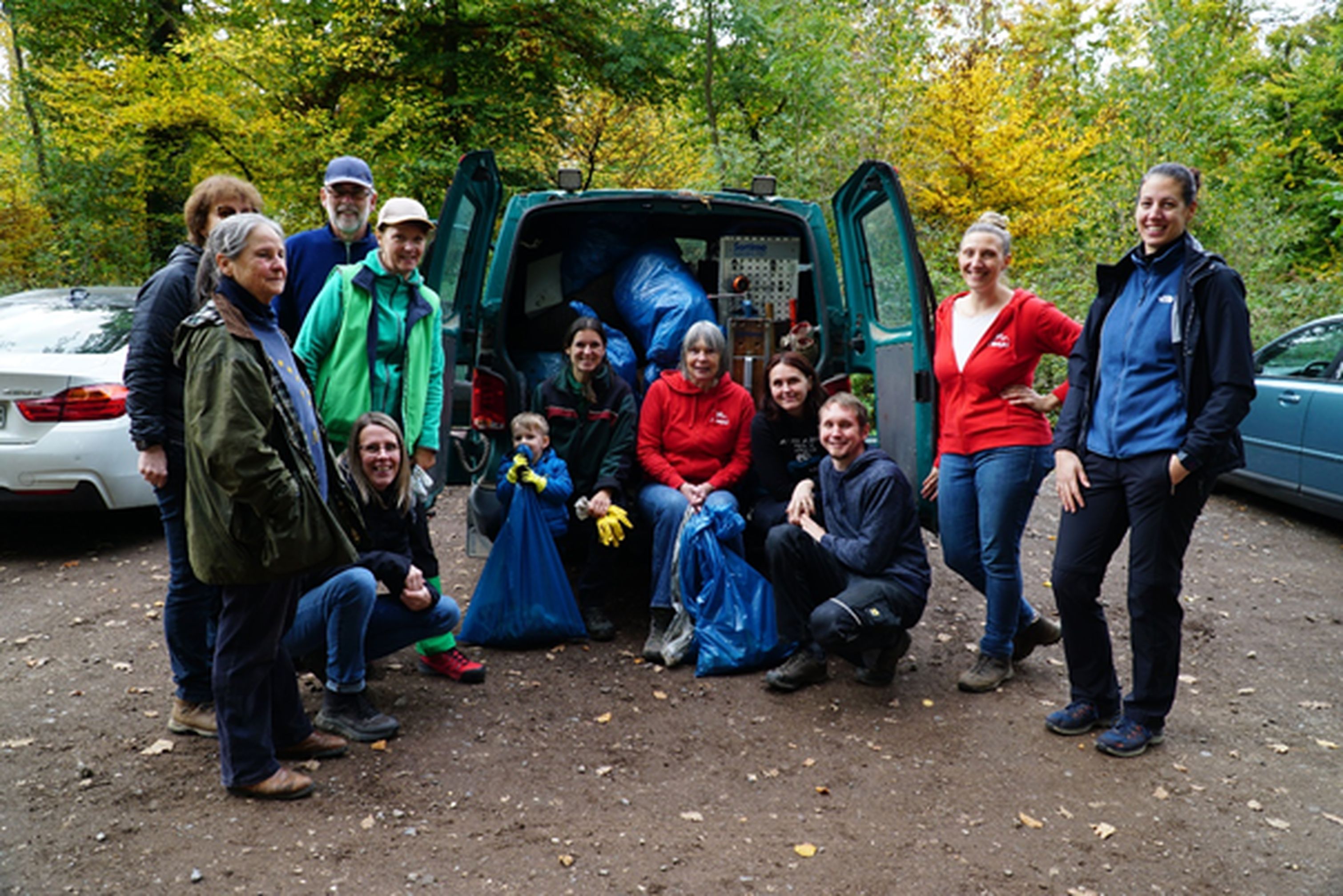 Eine größere Gruppe von Menschen steht bei einem Auto, dessen geöffneter Laderaum mit vielen Müllsäcken gefüllt ist.