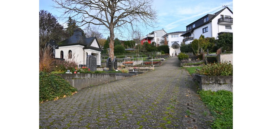 Ein gepflasterter Weg mit sichtbaren Schadstellen mit Gräbern an beiden Seiten. Im Hintergrund steht eine Trauerhalle.