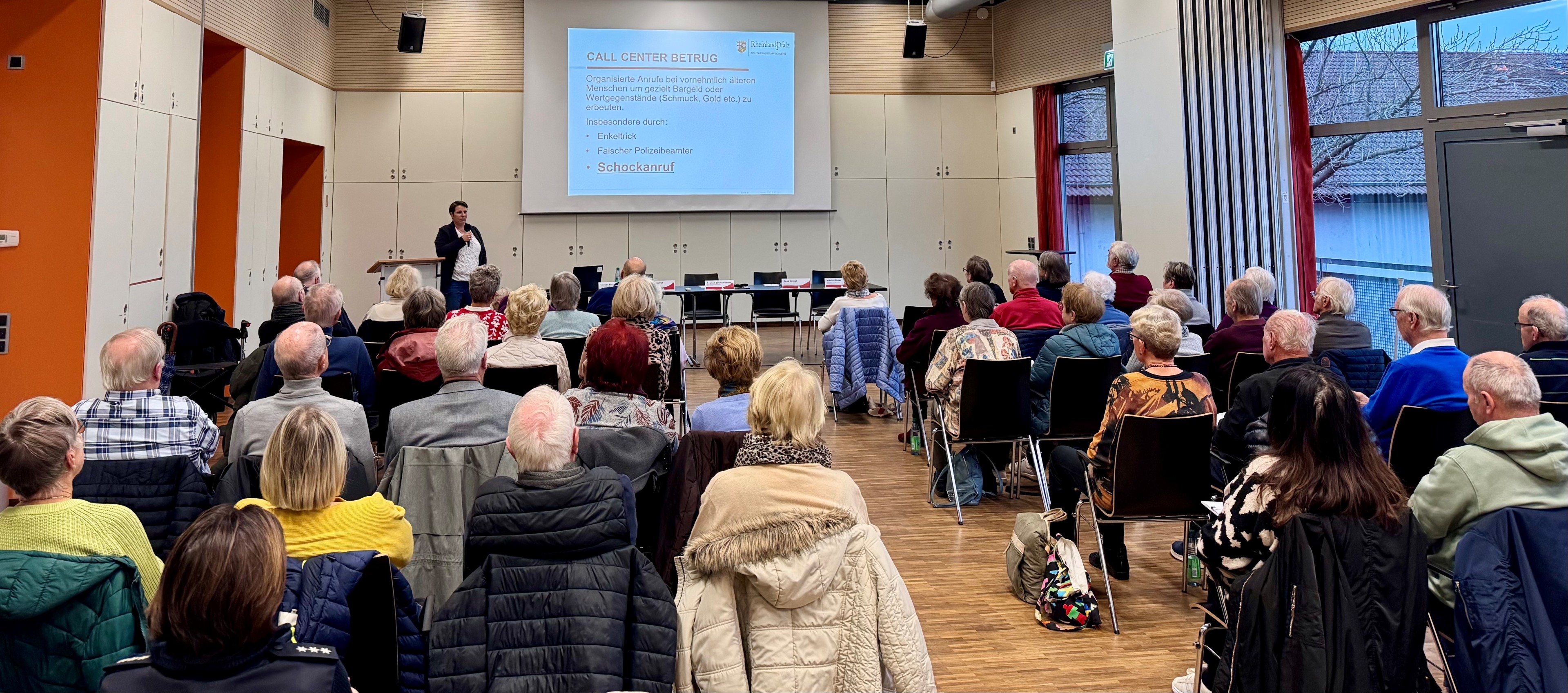 Viele Menschen in einem Saal. Vorne steht eine Rednerin.