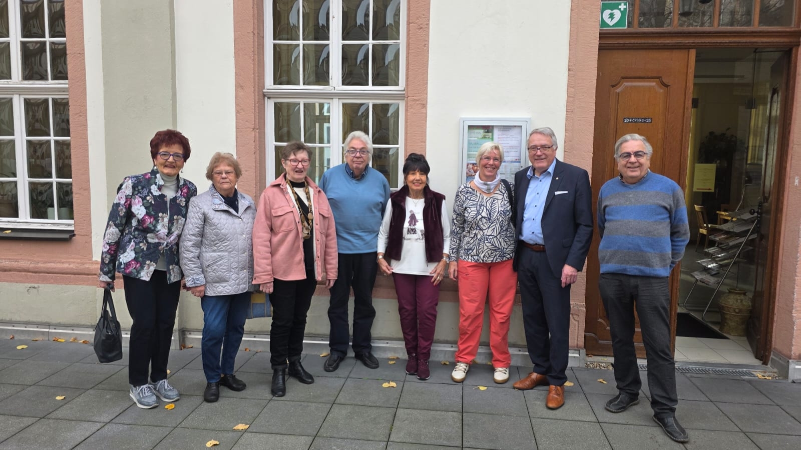 Vor einer Hauswand stehen eine Reihe von Menschen und Blicken in die Kamera.