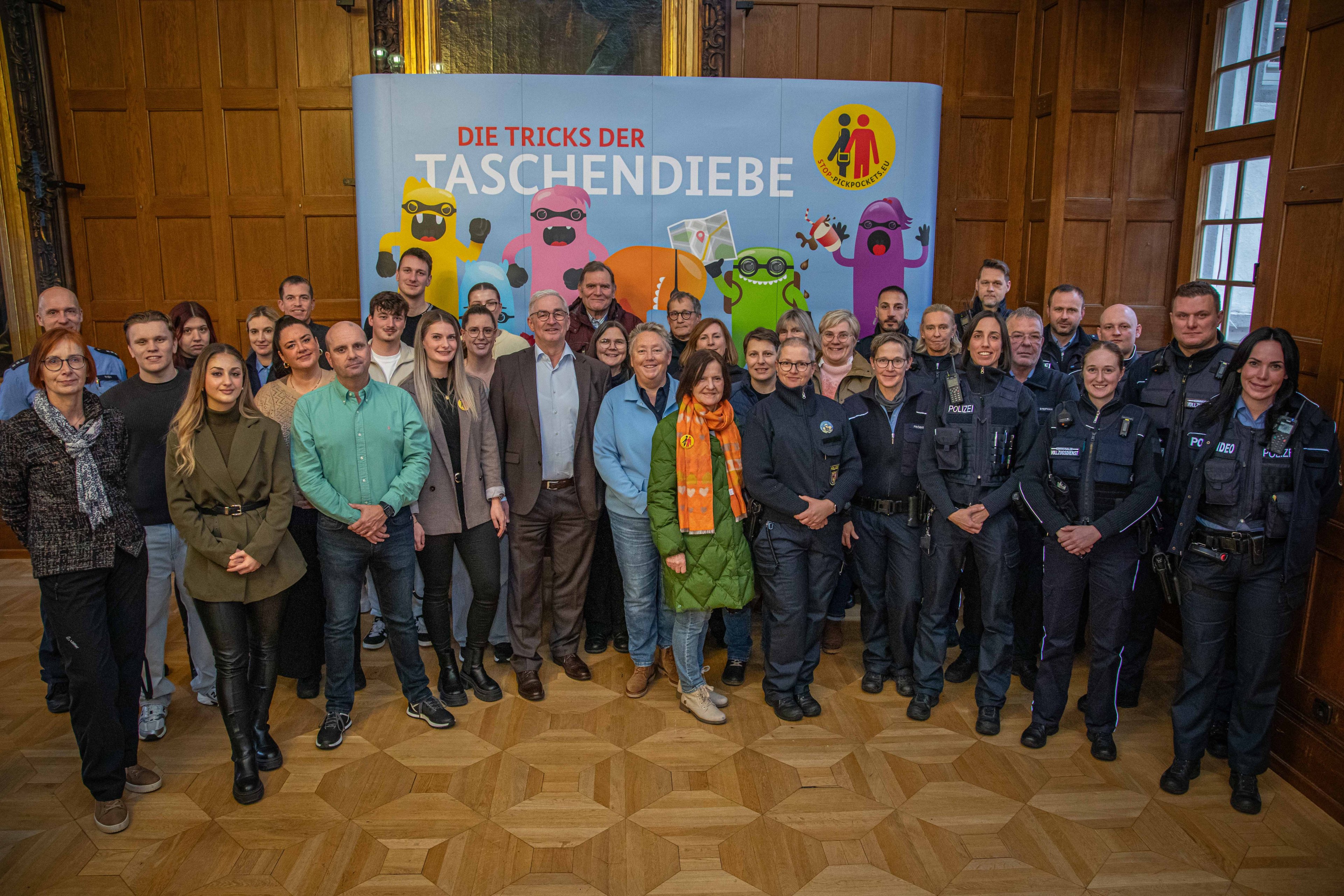 Viele Menschen, teils in Uniform, stehen in einem Saal vor goldgerahmten Portraitbildern.