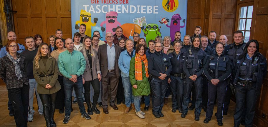 Viele Menschen, teils in Uniform, stehen in einem Saal vor goldgerahmten Portraitbildern.