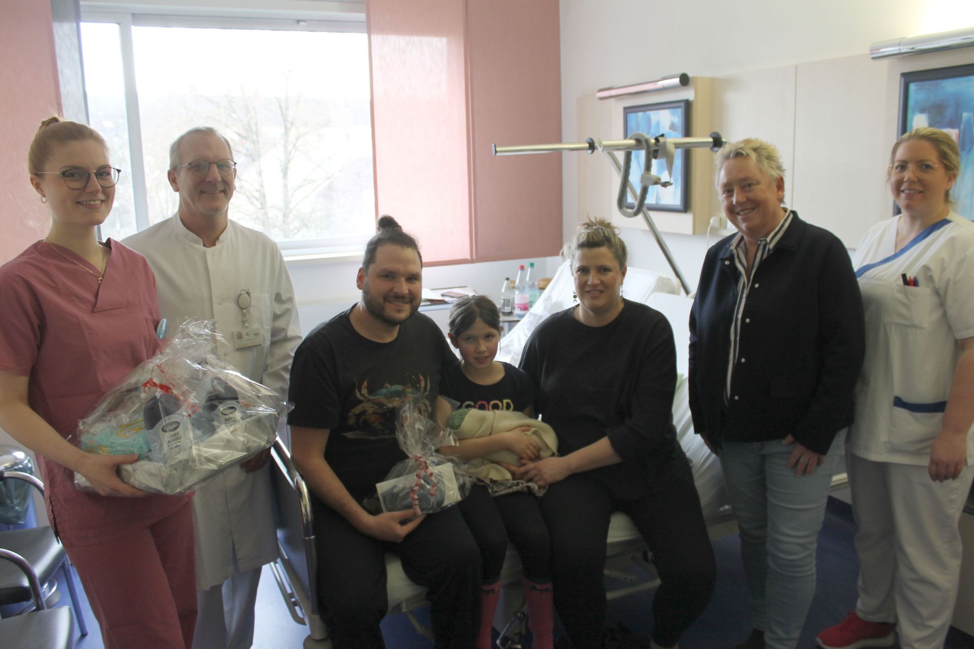 In einem Krankenhauszimmer sitzen eine vierköpfige Familie auf dem Bett und vier Erwachsene stehen neben ihnen.