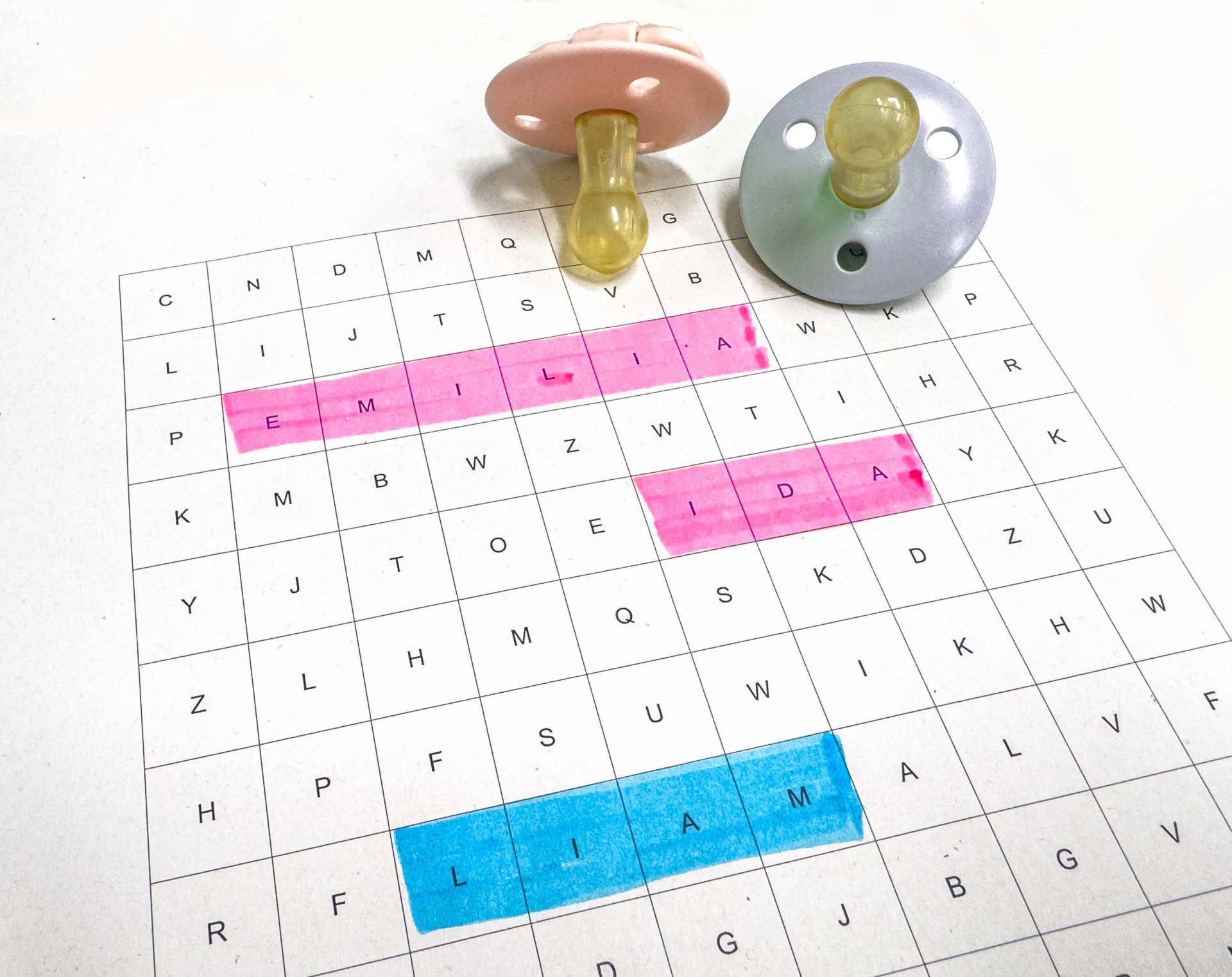 Zwei Schnuller liegen neben einem Wörterrätsel, in dem die Namen Emilia, Ida und Liam farblich markiert sind.