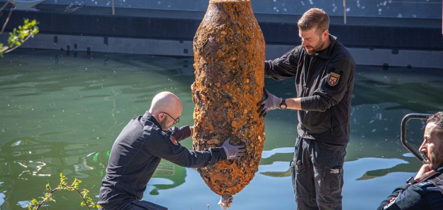 Zwei Männer halten eine Bombe, die an einem Kran hängt, im Gleichgewicht.