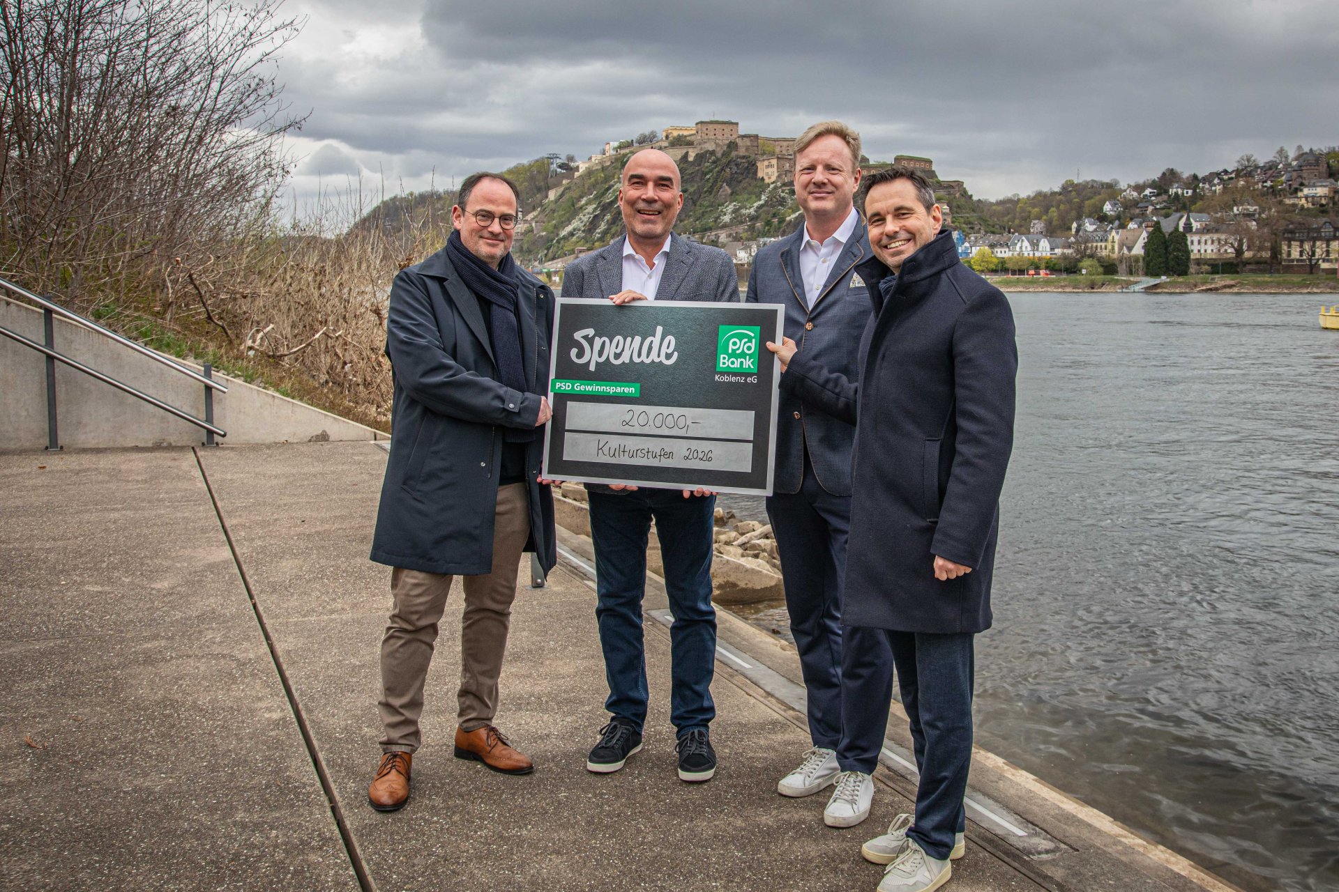 Vier Männer stehen mit einem Spendenscheck am Rheinufer.