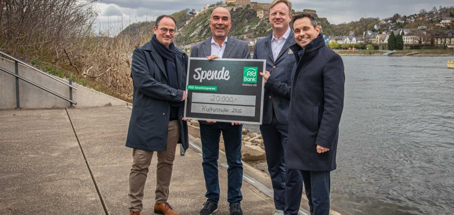 Vier Männer stehen mit einem Spendenscheck am Rheinufer.