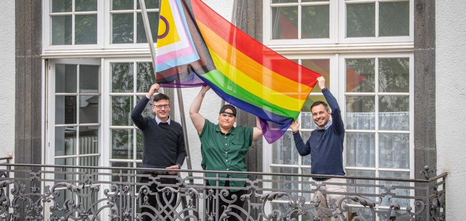Drei Personen stehen auf einem Balkon und halten eine Flagge in ihren Händen.