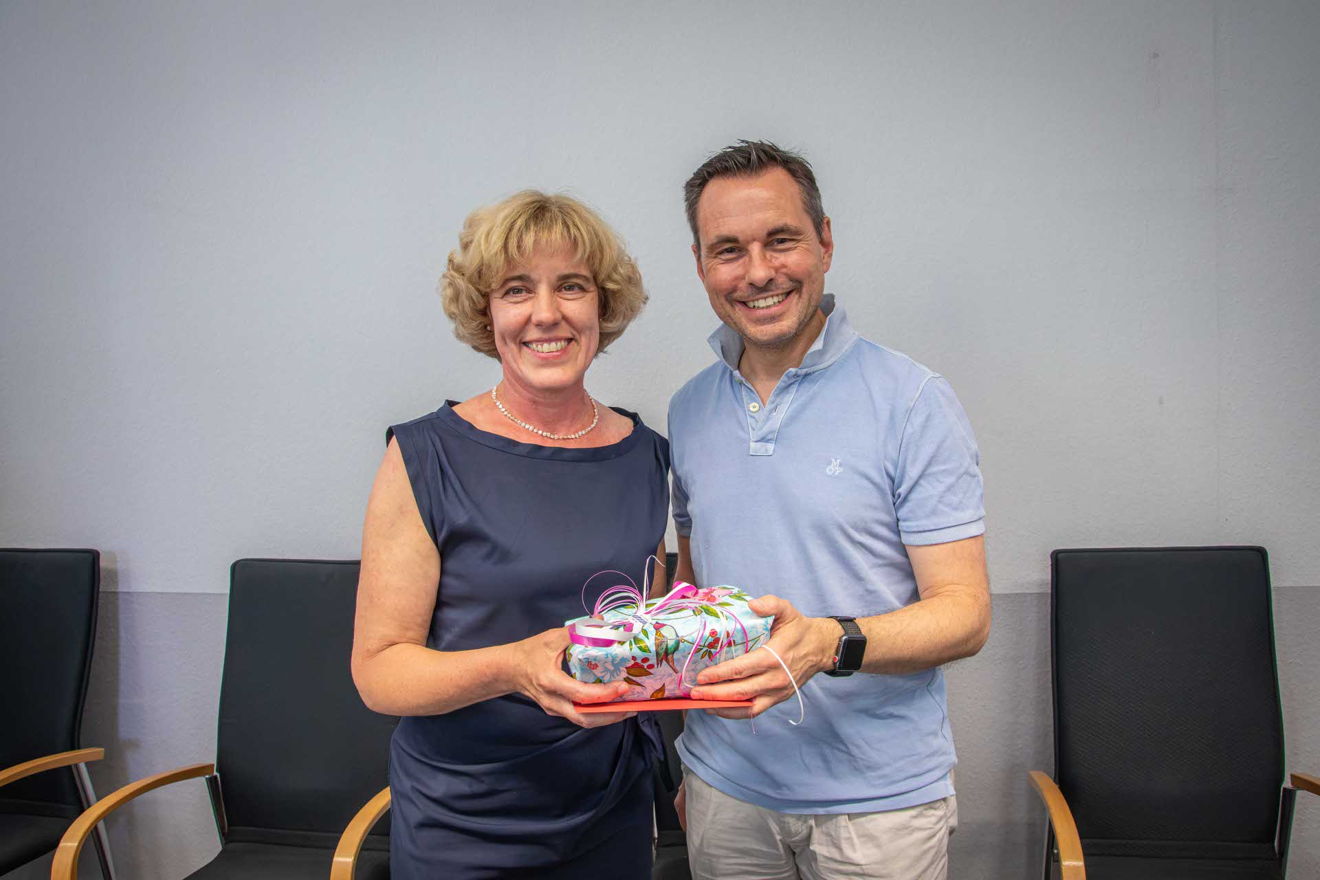 Eine Frau und ein Mann stehen mit einem Geschenk in der Hand vor einer Wand.