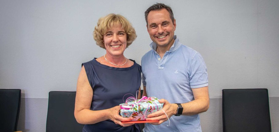 Eine Frau und ein Mann stehen mit einem Geschenk in der Hand vor einer Wand. Eine Frau und ein Mann stehen mit einem Geschenk in der Hand vor einer Wand.