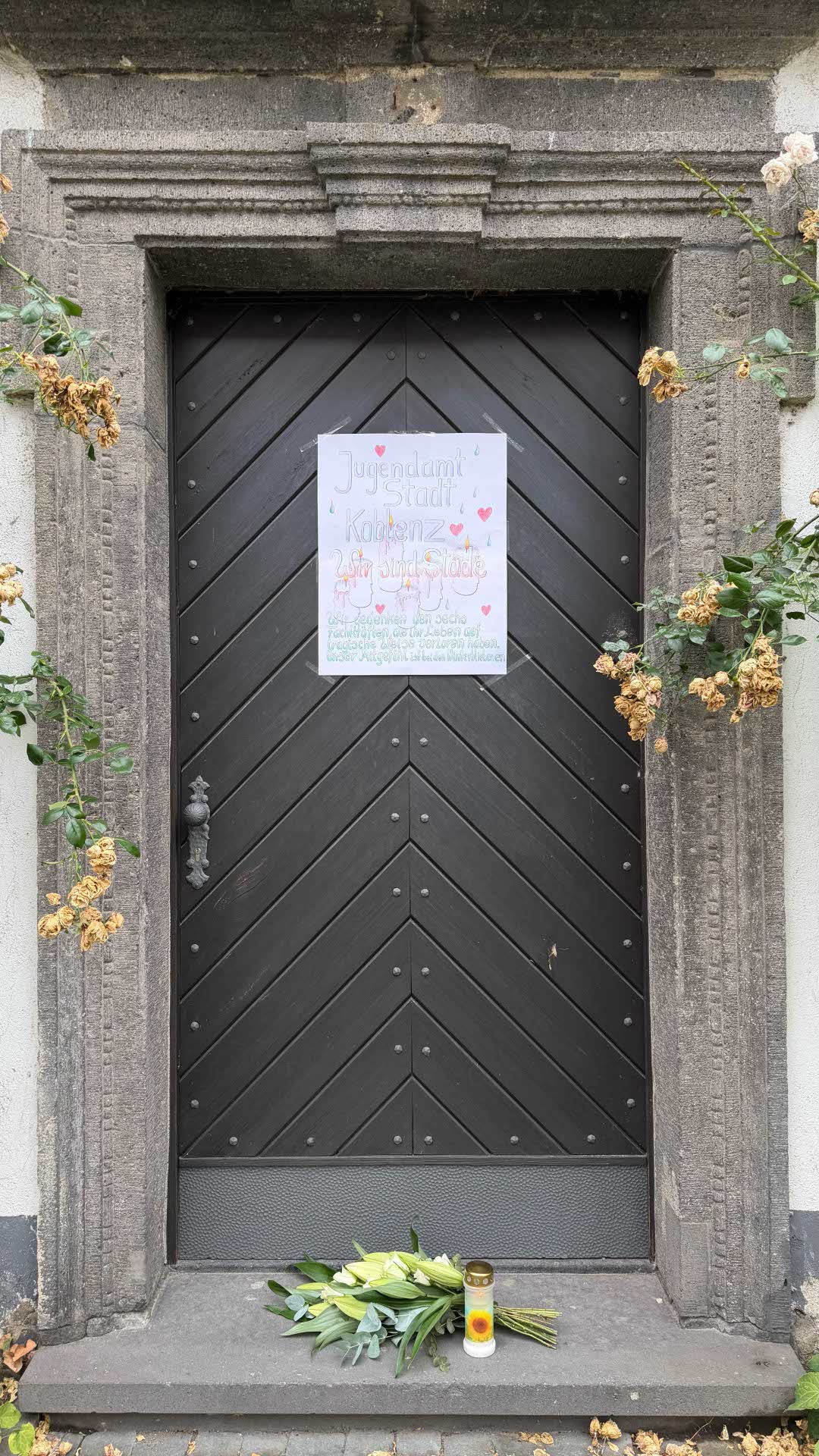 Ein Plakat hängt an einer Tür. Davor ein Blumenstrauß und eine Kerze.