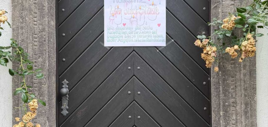 Ein Plakat hängt an einer Tür. Davor ein Blumenstrauß und eine Kerze.