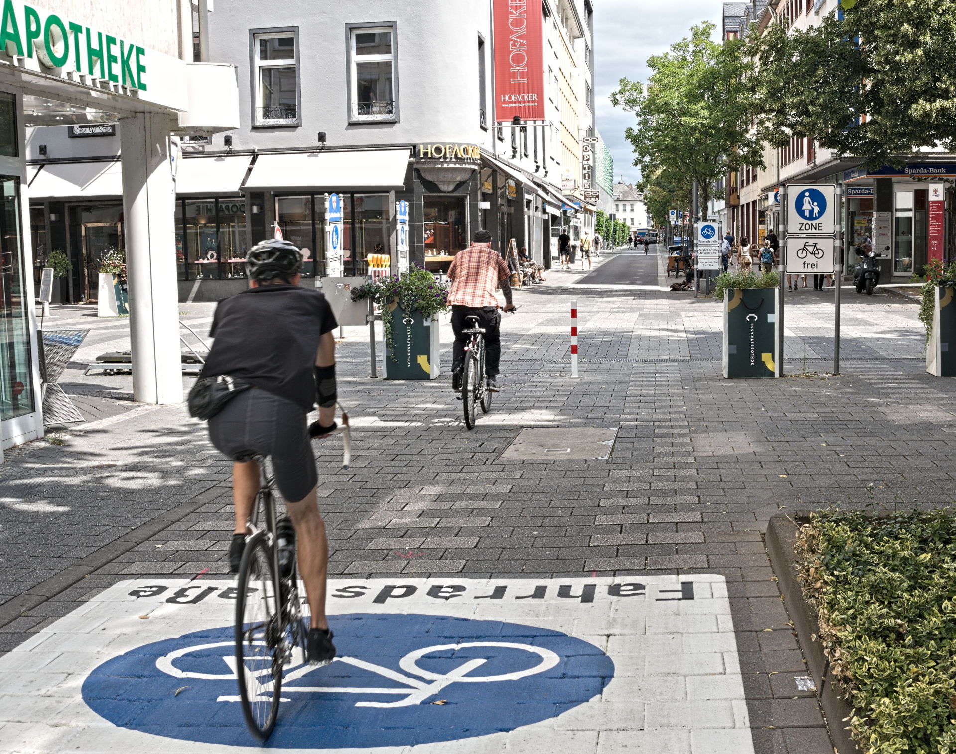 Zwei Radfahrer fahren durch eine Straße. In der Straßenmitte steht ein rot-weißer Roller.