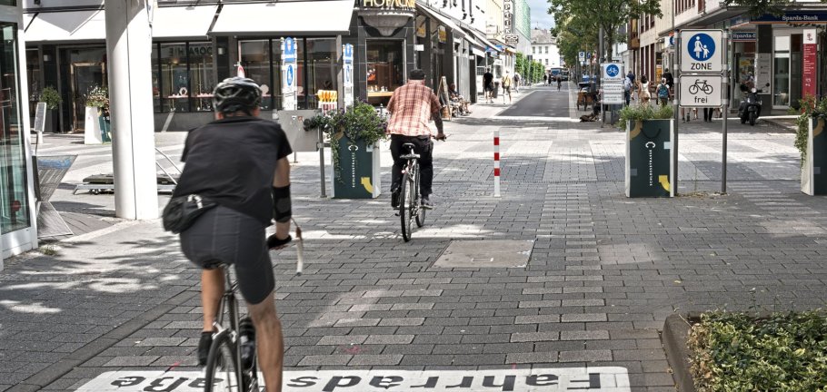 Zwei Radfahrer fahren durch eine Straße. In der Straßenmitte steht ein rot-weißer Roller.