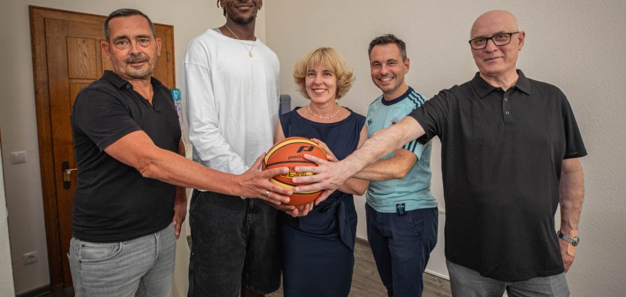 Fünf Personen fassen an einen Basketball und schauen in eine Kamera. Fünf Personen fassen an einen Basketball und schauen in eine Kamera.