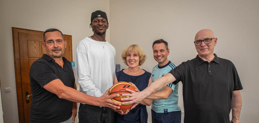 Fünf Personen fassen an einen Basketball und schauen in eine Kamera. Fünf Personen fassen an einen Basketball und schauen in eine Kamera.