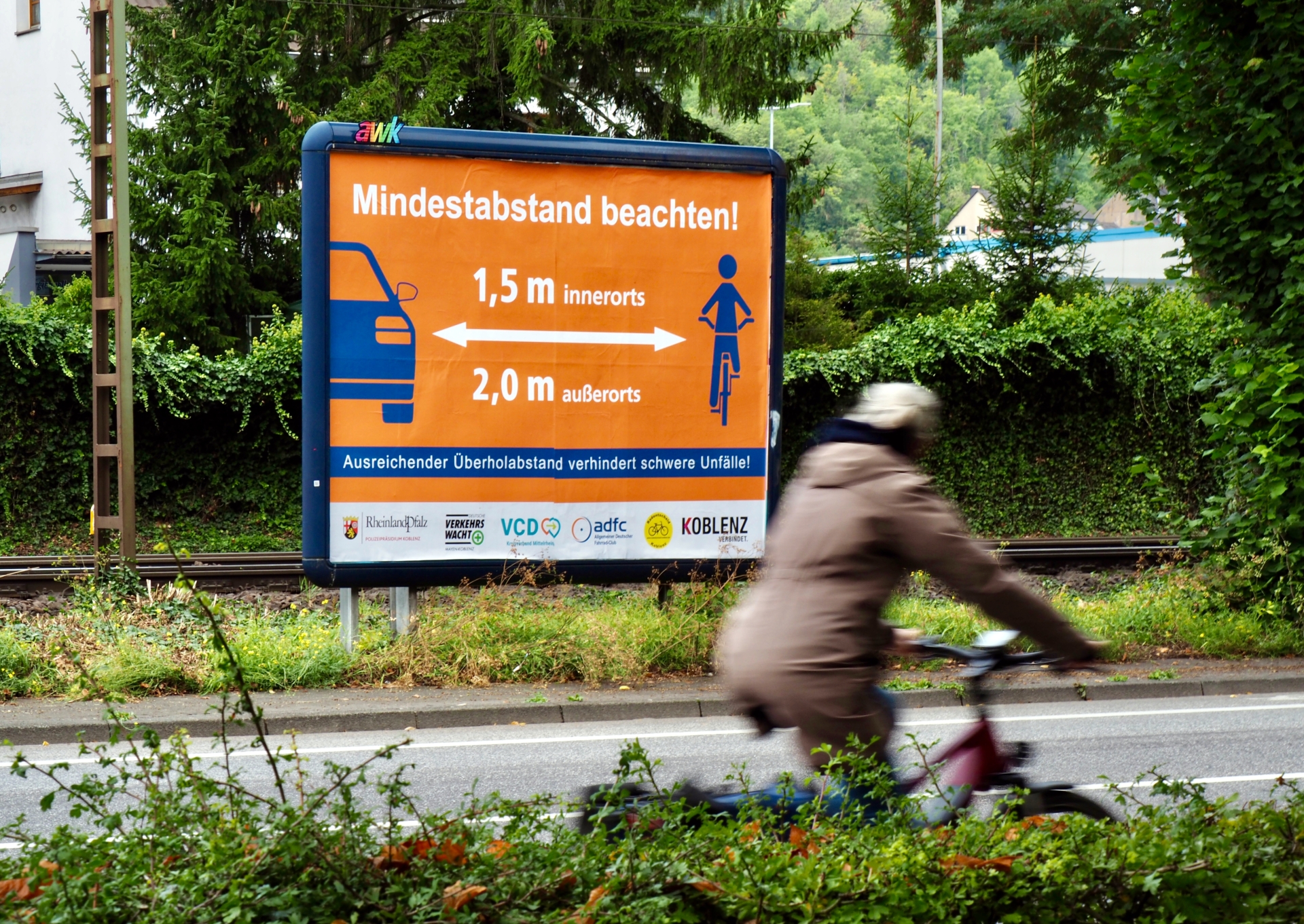 Eine Fahrradfahrerin fährt an einem Plakat vorbei. Auf dem Plakat wird auf einen Mindestabstand zwischen Fahrzeugen und Fahrrädern hingewiesen.