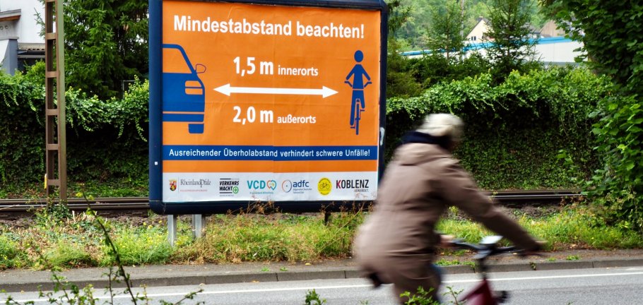 Eine Fahrradfahrerin fährt an einem Plakat vorbei. Auf dem Plakat wird auf einen Mindestabstand zwischen Fahrzeugen und Fahrrädern hingewiesen. Eine Fahrradfahrerin fährt an einem Plakat vorbei. Auf dem Plakat wird auf einen Mindestabstand zwischen Fahrzeugen und Fahrrädern hingewiesen.