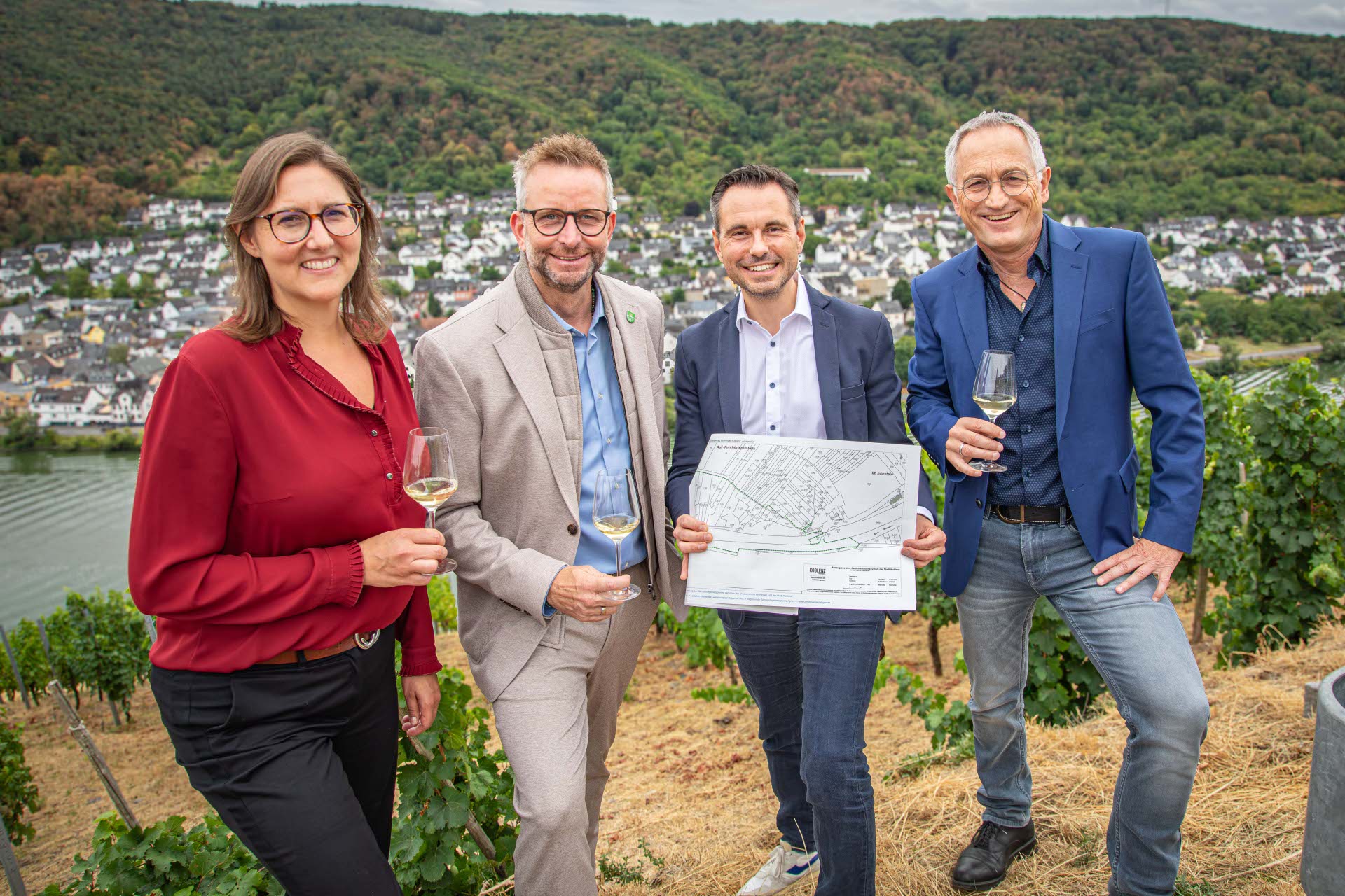 Eine Frau und drei Männer stehen mit Weingläser und einem Flurkarte in einem Weinberg.