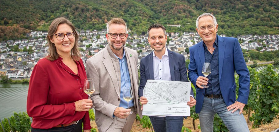 Eine Frau und drei Männer stehen mit Weingläser und einem Flurkarte in einem Weinberg.