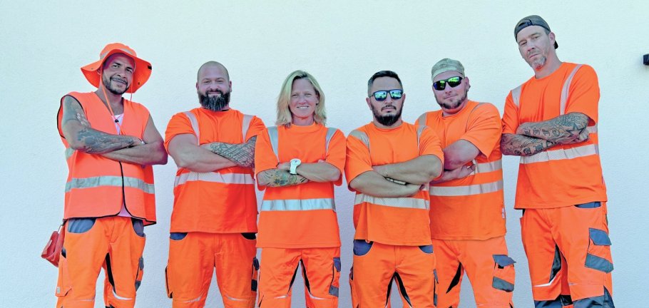 Fünf Männer und eine Frau stehen in orangenen Arbeitsklamotten vor einer Wand. Fünf Männer und eine Frau stehen in orangenen Arbeitsklamotten vor einer Wand.