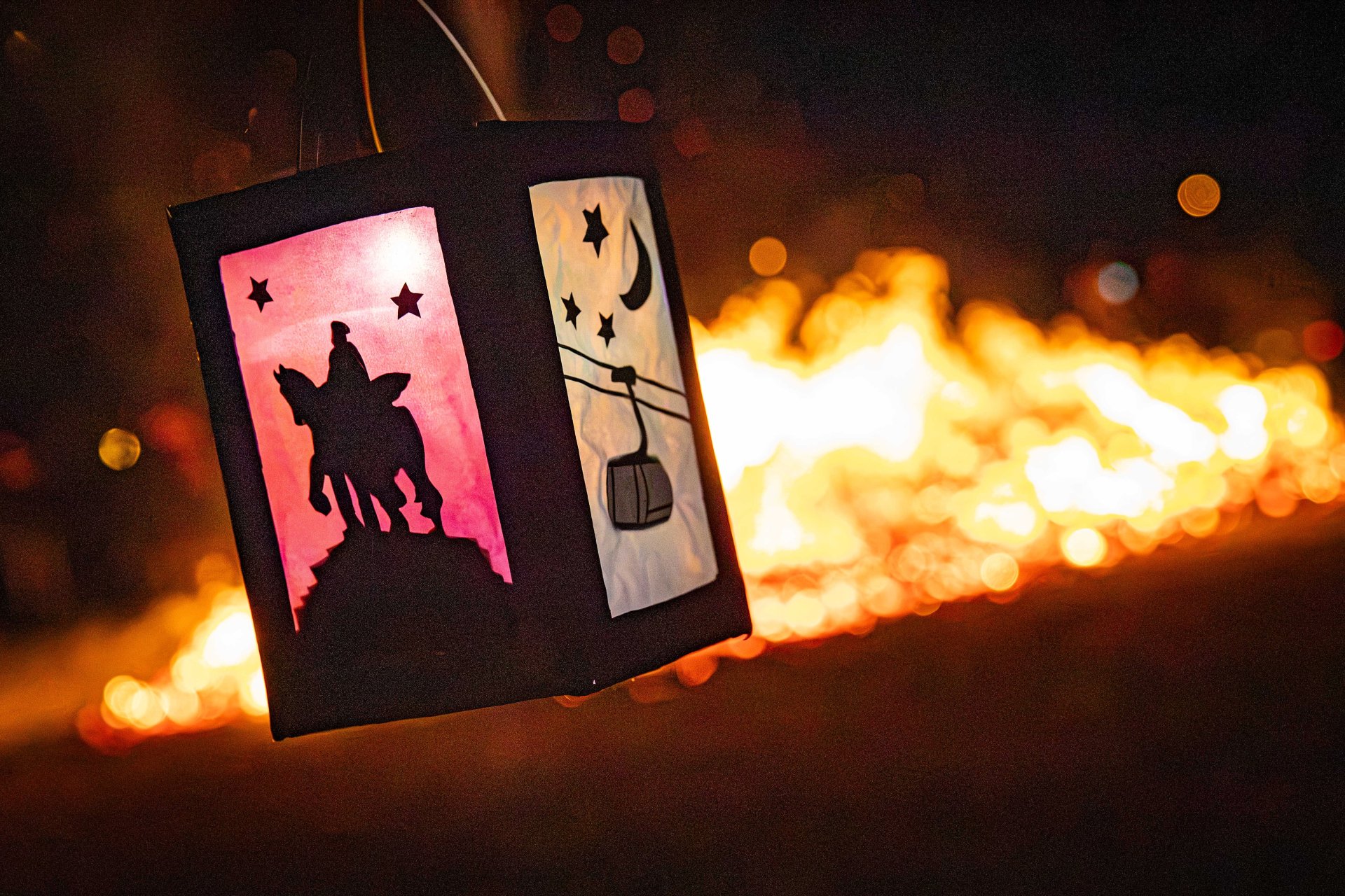 Eine Laterne leuchtet im Vordergrund, während im Hintergrund ein Feuer zu sehen ist.